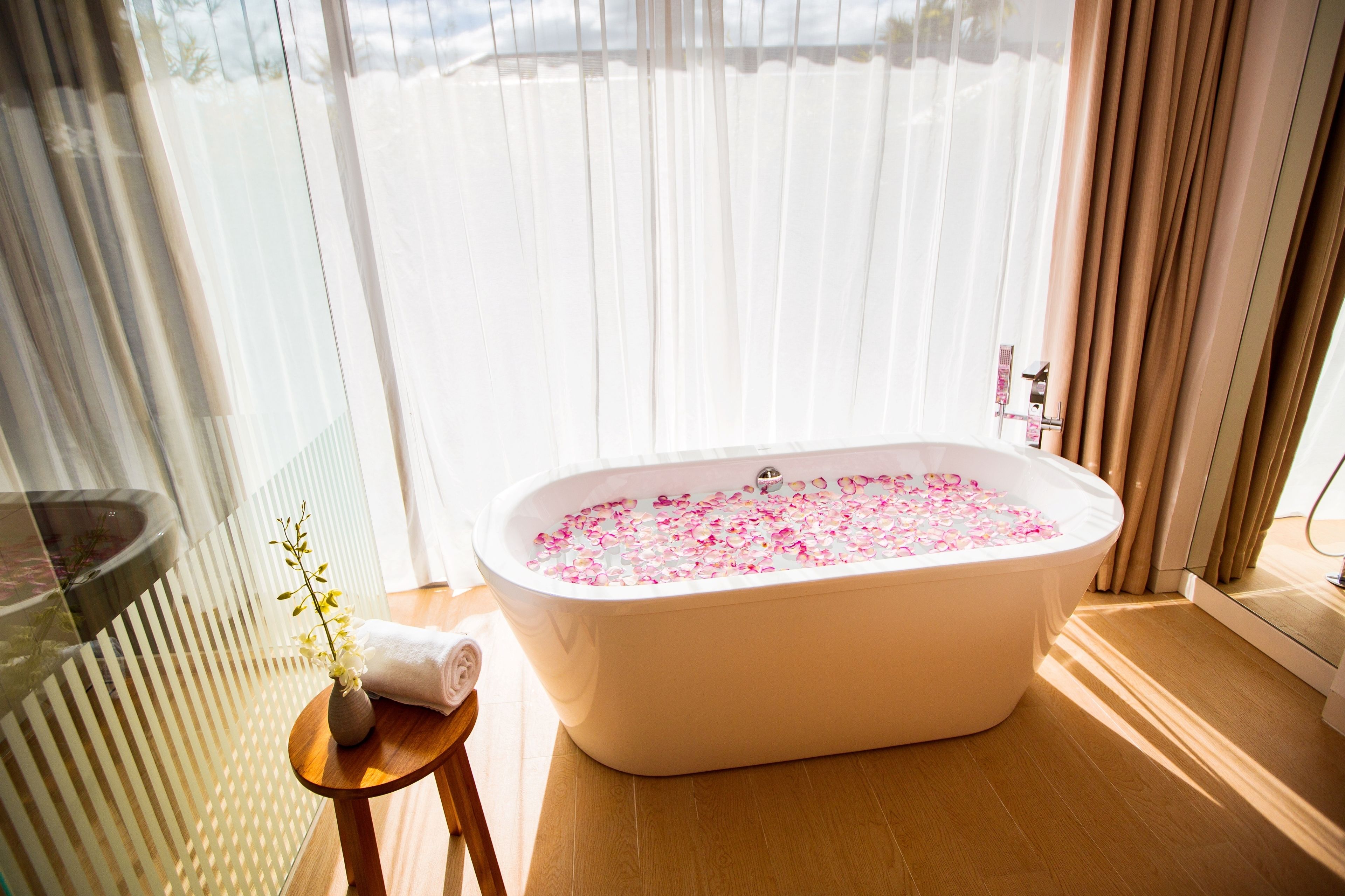 A bathroom with a freestanding bathtub filled with rose petals, a small wooden stool with a vase, and a glass shower enclosure.