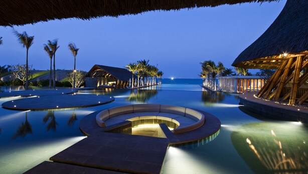 A serene infinity pool with a circular seating area, surrounded by palm trees and thatched-roof structures, reflecting a tranquil evening setting. - Naman Retreat (Da Nang)