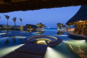 Una piscina infinita serena con una zona de estar circular, rodeada de palmeras y construcciones de techo de paja, que refleja un ambiente vespertino tranquilo.