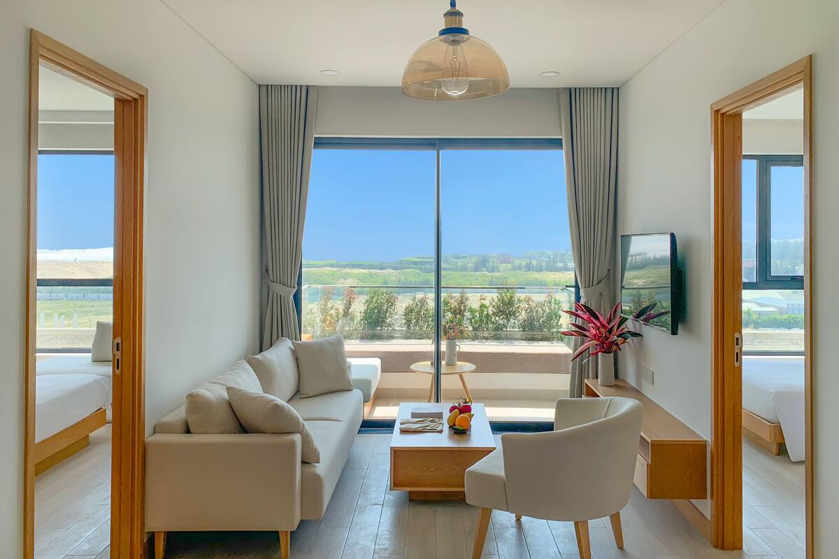 Un salon moderne avec un canapé, une table basse et un téléviseur, de grandes fenêtres offrant une vue sur la verdure et un ciel dégagé.