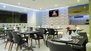 A modern restaurant with black chairs and white table settings, a TV on the wall, and a patterned wall. - The Life Hotels (Surabaya)