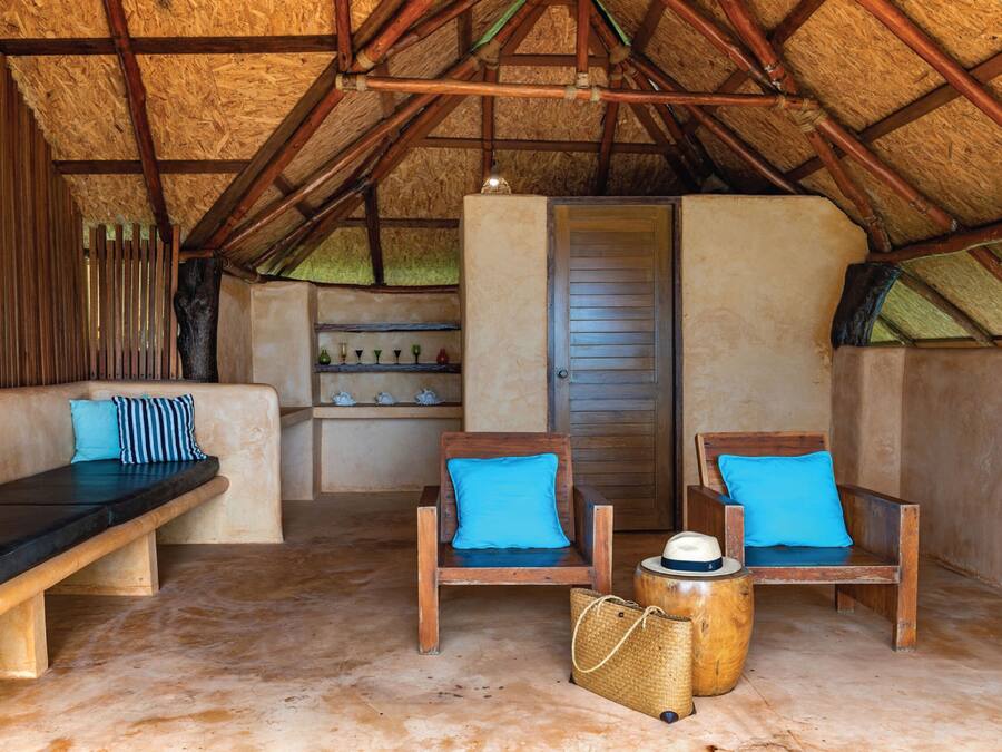 A cozy room with a thatched roof, wooden benches with blue cushions, a bench with a black cushion, and a woven basket on the floor.