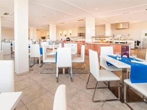 Un comedor moderno con mesas y sillas blancas, una isla central con cocina y un diseño luminoso y espacioso.