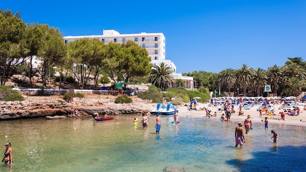 A beach with clear water, people swimming, and a white building in the background. - Globales Cala'n Blanes (Ciutadella de Menorca)