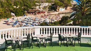 A terrace with outdoor seating and a view of a beach with umbrellas and people. - Globales Cala'n Blanes (Ciutadella de Menorca)