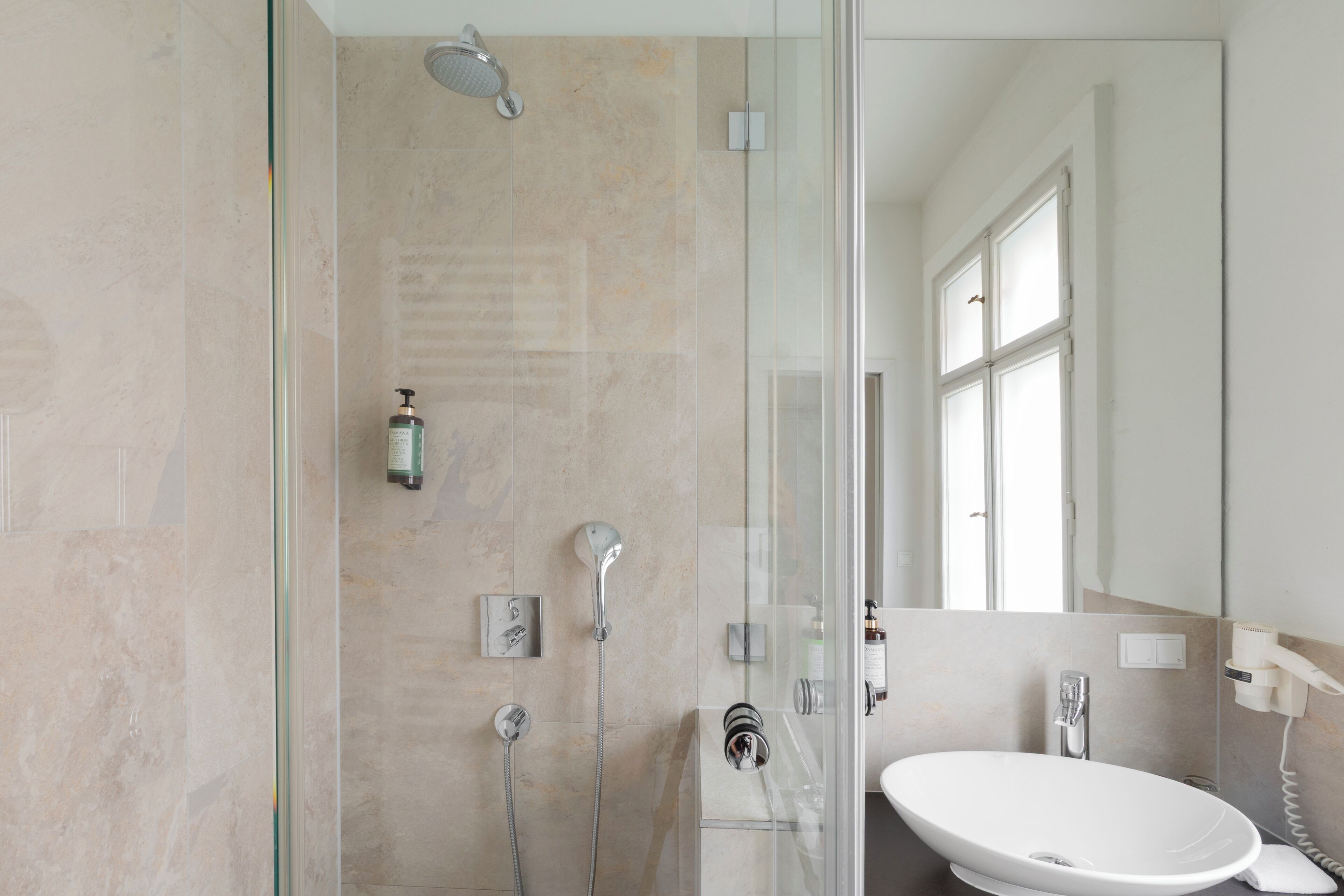 Un baño moderno con cabina de ducha de vidrio, un lavabo blanco y un espejo que refleja una ventana.