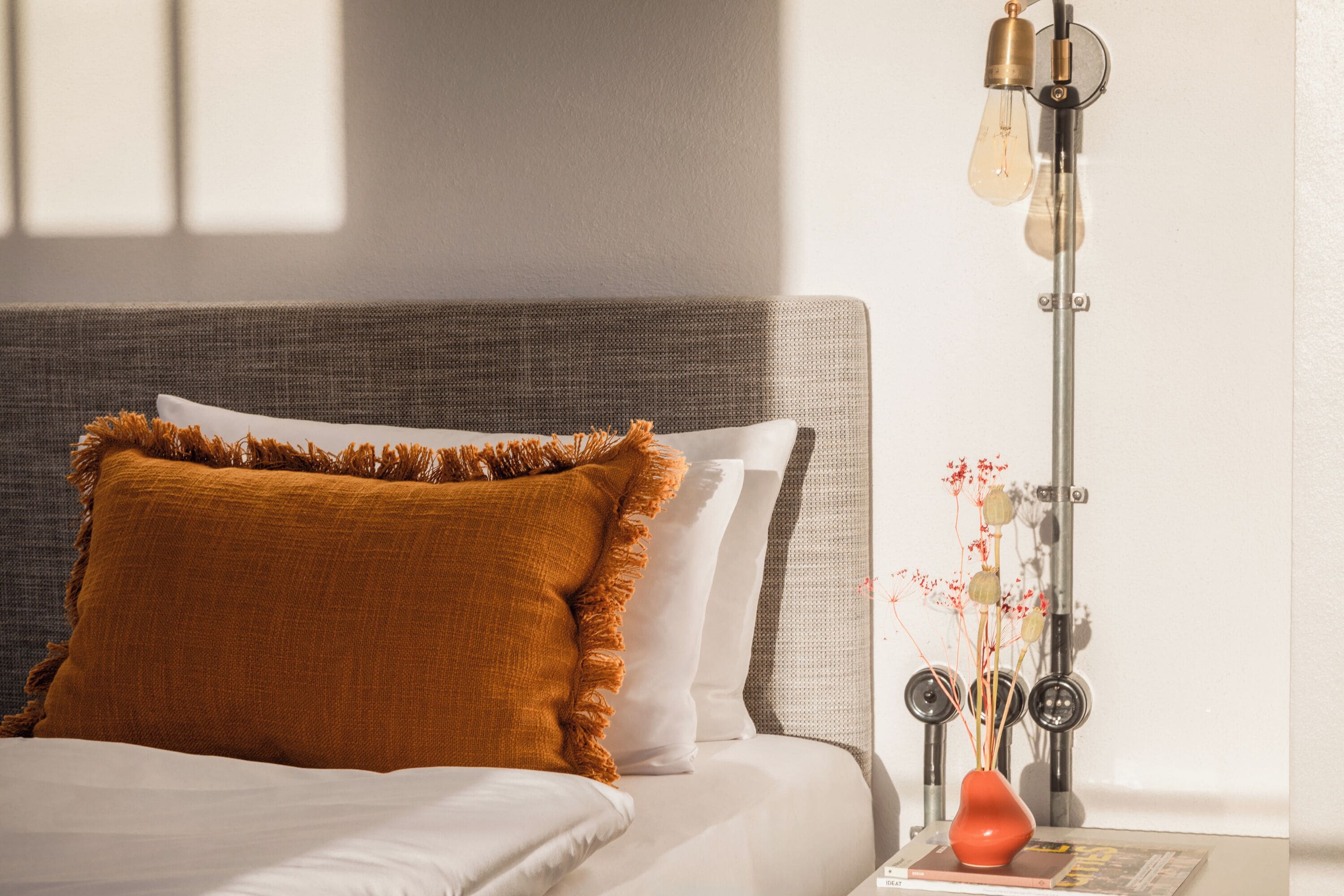 A neatly made bed with a textured headboard, a bedside table with a vase of dried flowers, and a modern floor lamp.