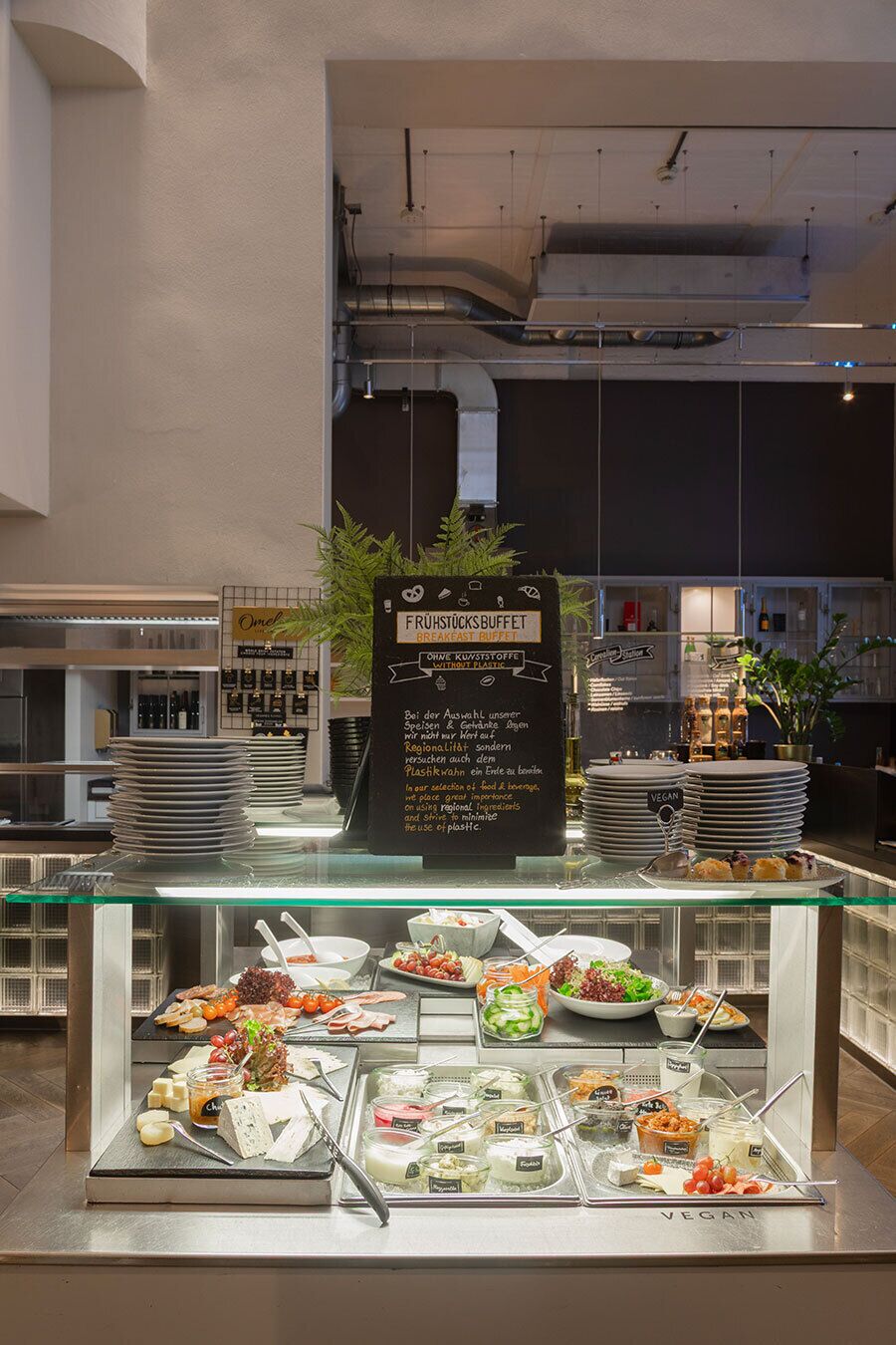 A breakfast buffet area with various food options, including vegan selections, in a modern hotel setting.