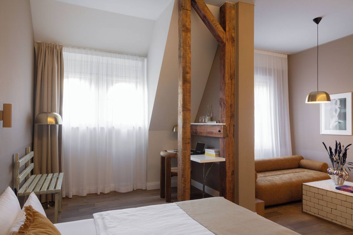 A modern hotel room with a bed, a sofa, a desk, and a large window with curtains.