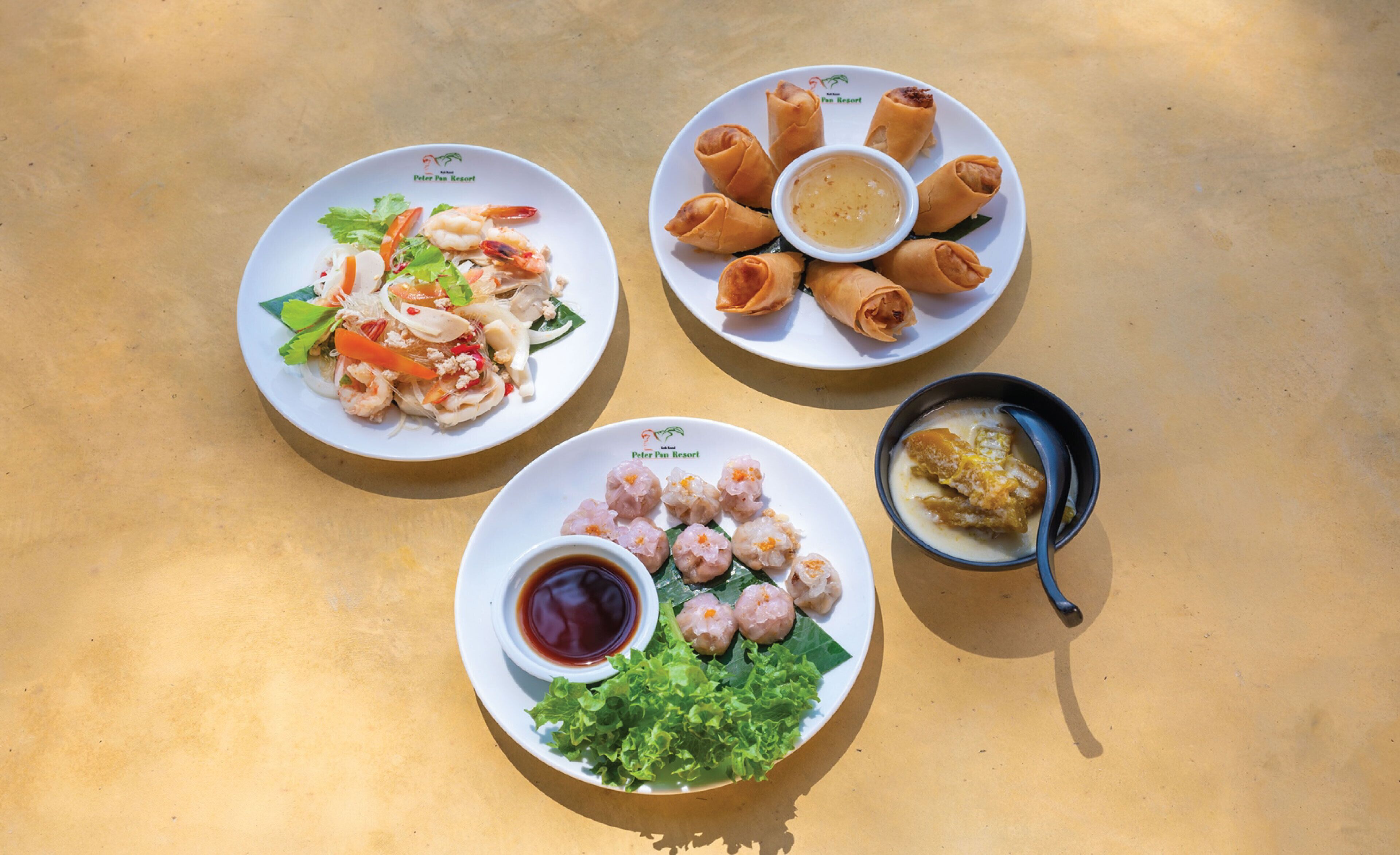A table with three plates of food: one with a shrimp salad, one with spring rolls and dipping sauce, and one with shrimp dumplings and dipping sauce.