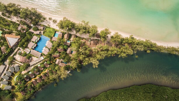 An aerial view of a coastal resort with a swimming pool, beachfront villas, and lush greenery.