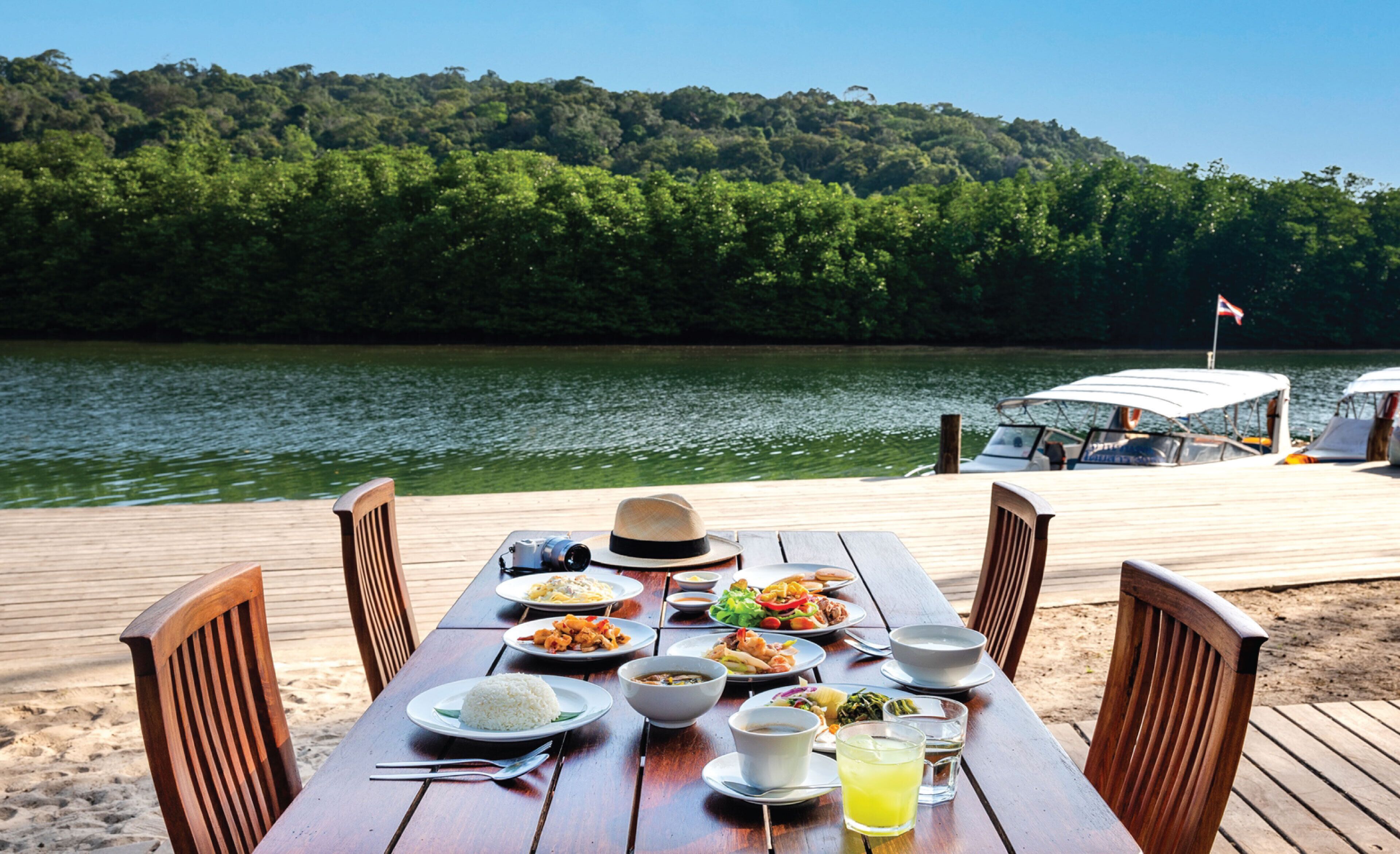 Una mesa de madera con platos de comida, un sombrero y una bebida, sobre un muelle con un bote y colinas boscosas al fondo.