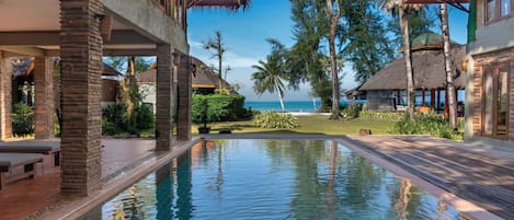 A resort pool area with clear blue water, surrounded by palm trees and thatched-roof structures.