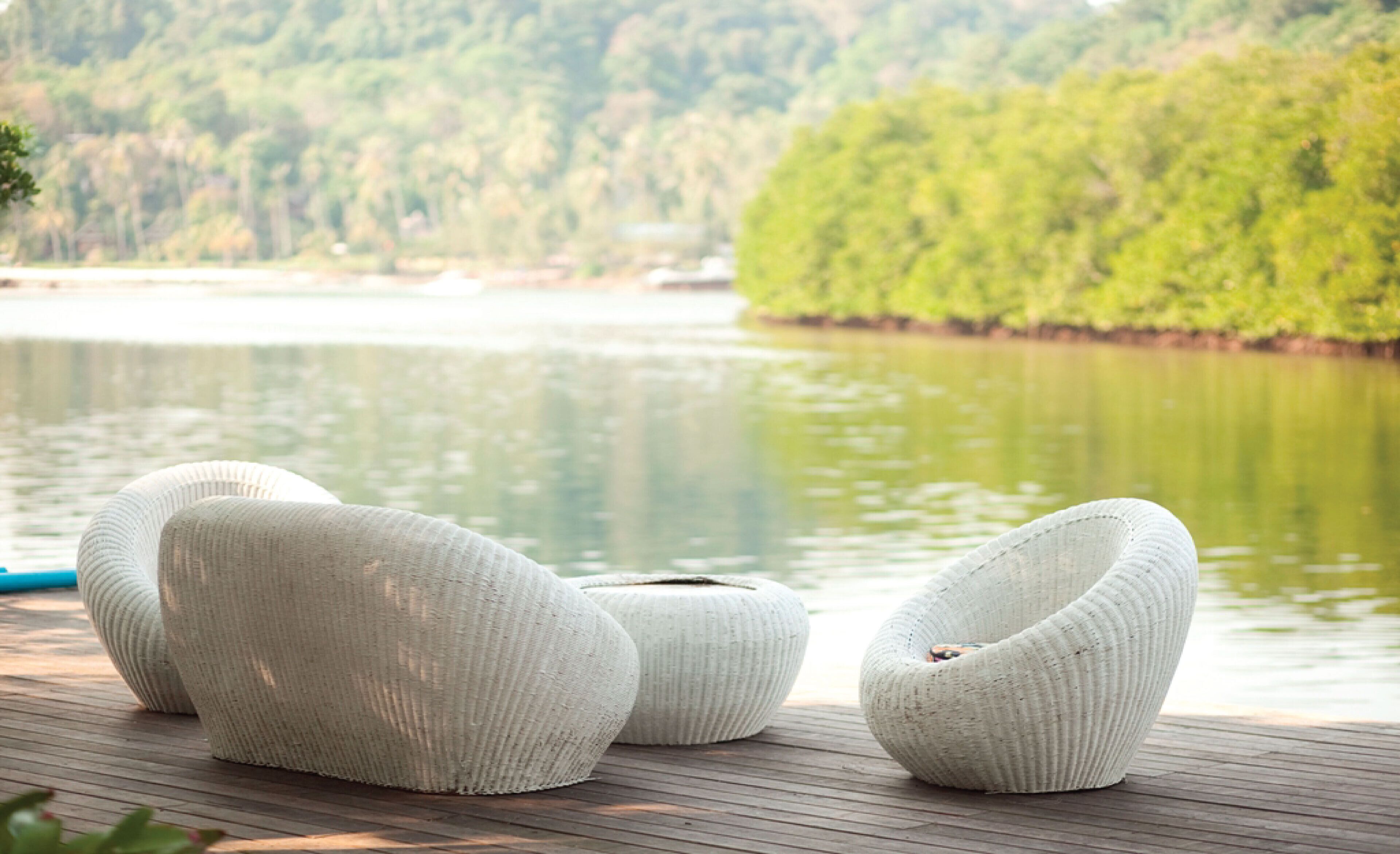 Three white, ribbed, woven chairs on a wooden deck by a calm lake with a forested hillside in the background.