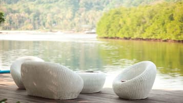 Three white, ribbed, woven chairs on a wooden deck by a calm lake with a forested hillside in the background.