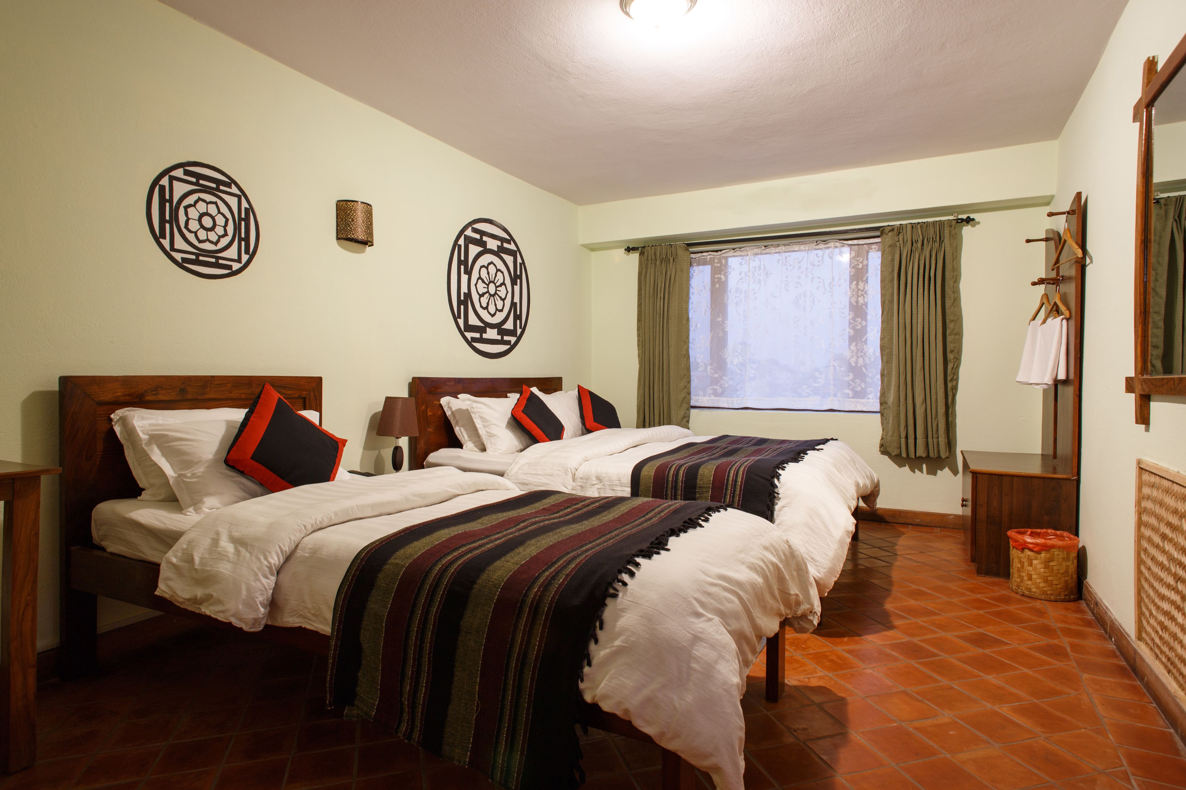 A bedroom with two beds, a window with curtains, a wall-mounted lamp, and a mirror.