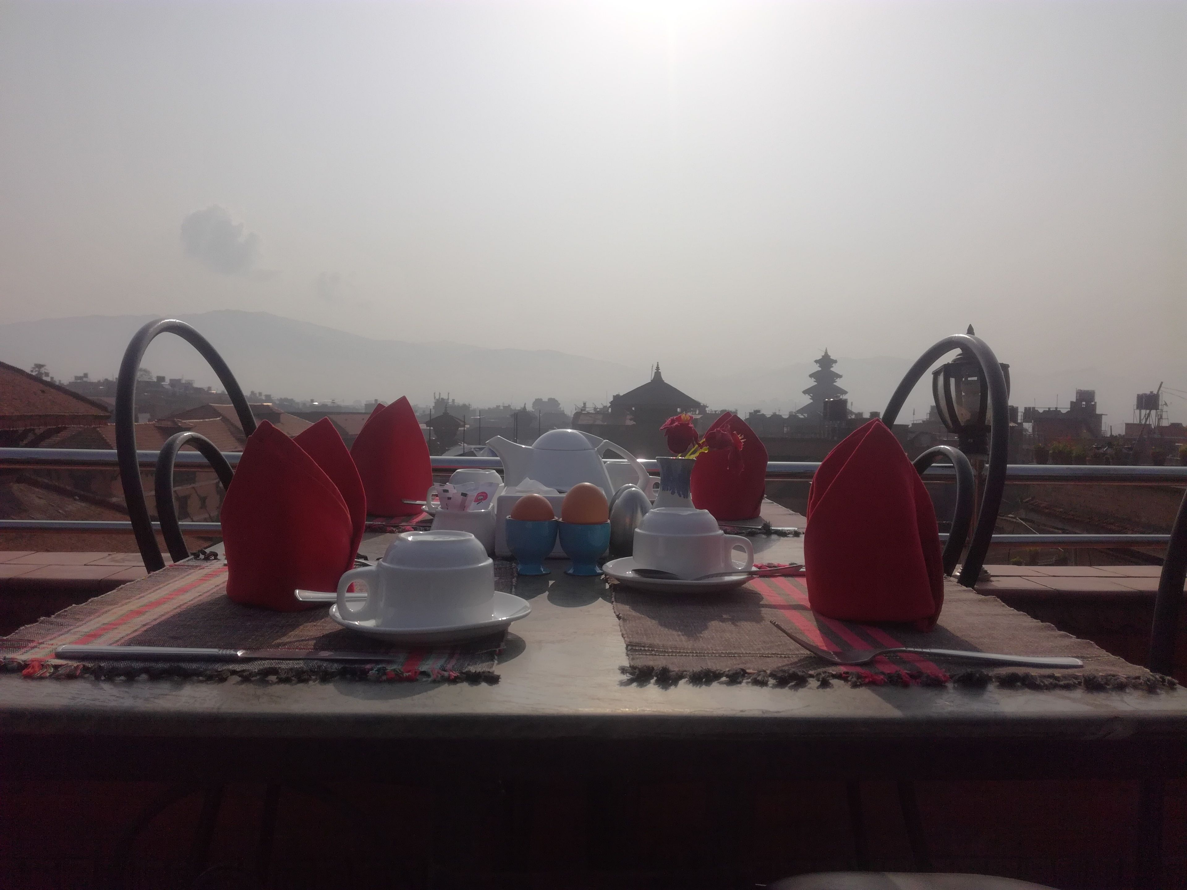 A dining table set with plates, cups, and napkins, overlooking a mountainous landscape with traditional architecture.