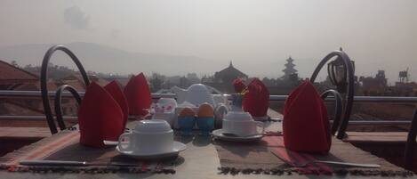 A dining table set with plates, cups, and napkins, overlooking a mountainous landscape with traditional architecture.