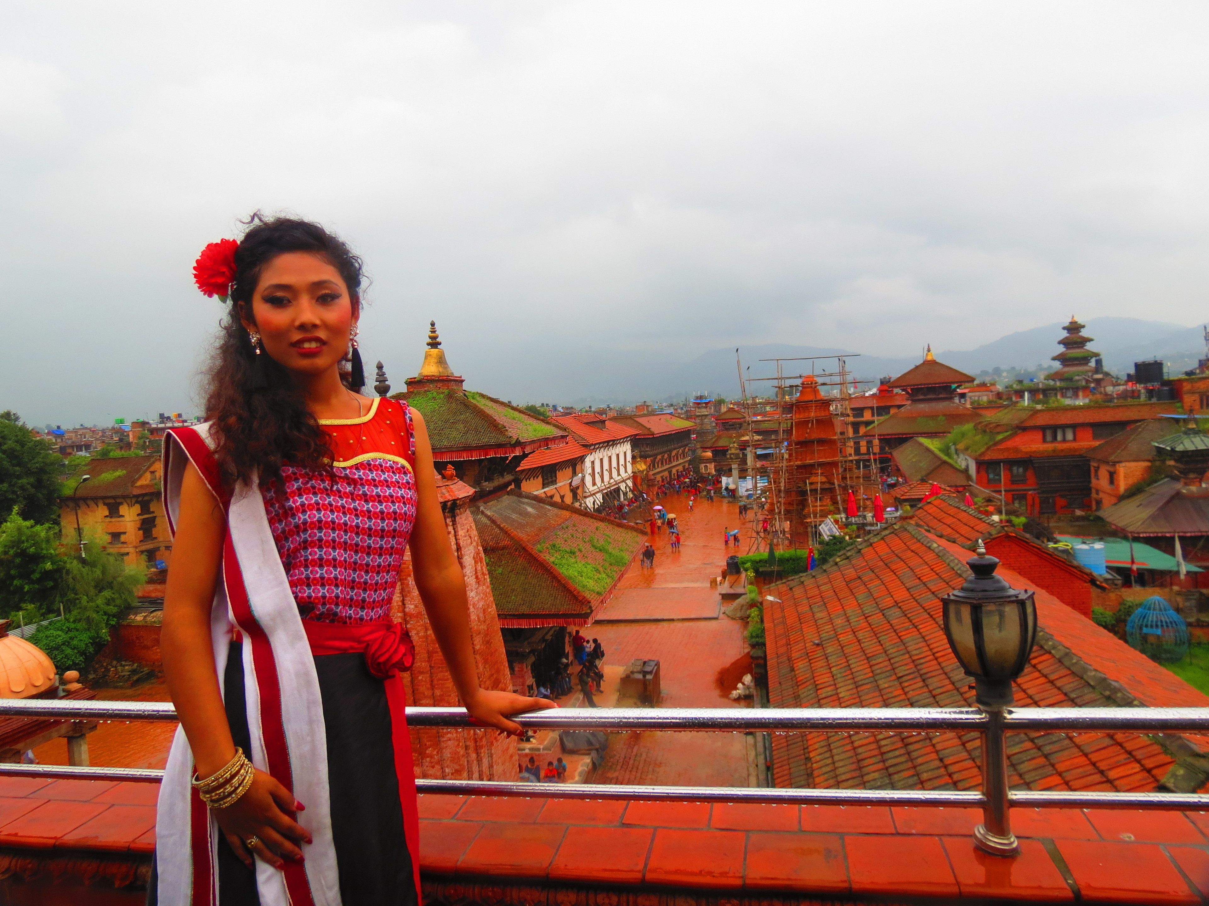 A person standing on a balcony overlooking a traditional town with red-roofed buildings and a temple.