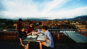 Un grupo de personas cenando en una mesa al aire libre con vistas a un paisaje urbano y montañas al fondo.