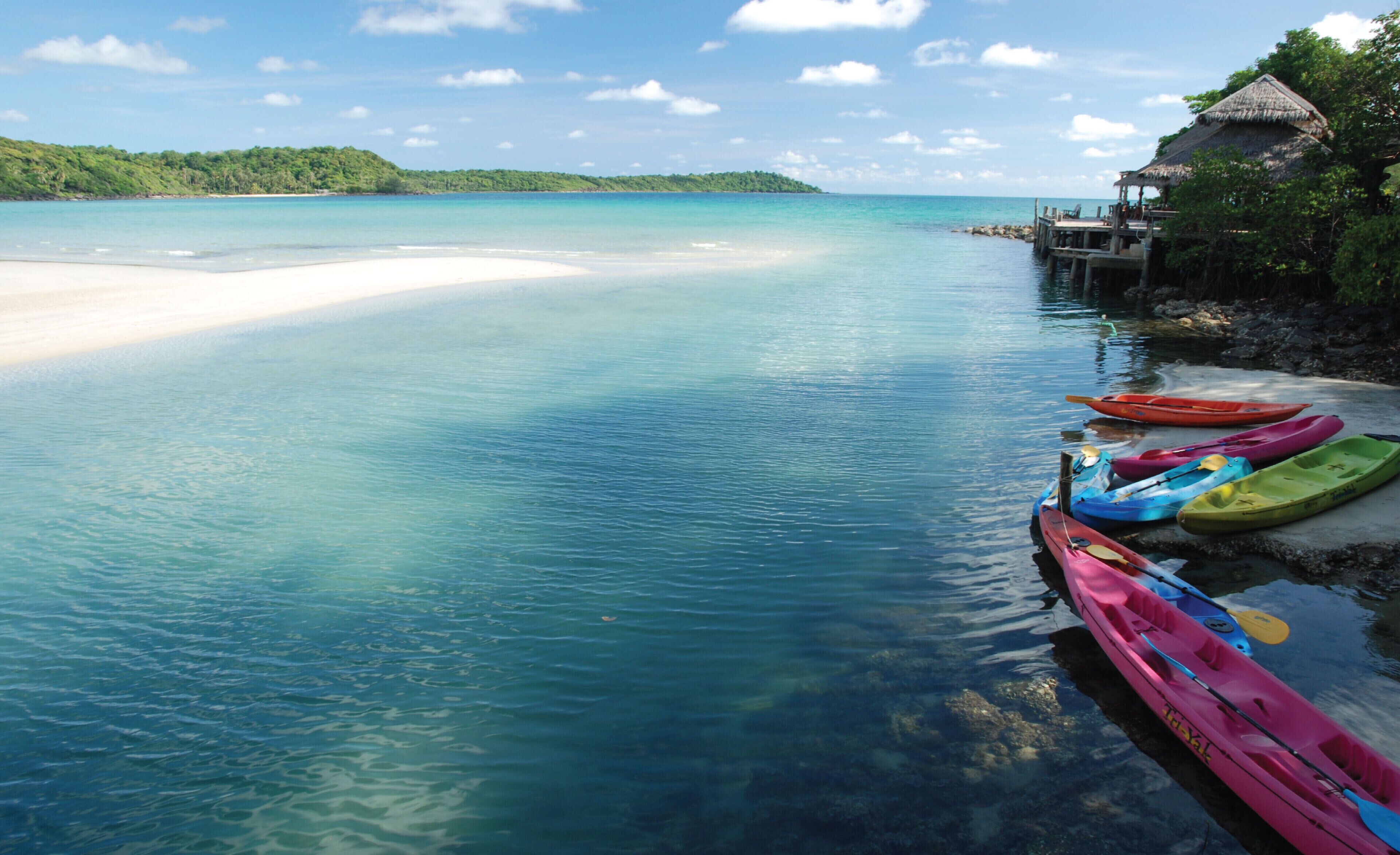 Una playa con agua turquesa cristalina, una orilla de arena blanca y varios kayaks de colores alineados a la derecha.