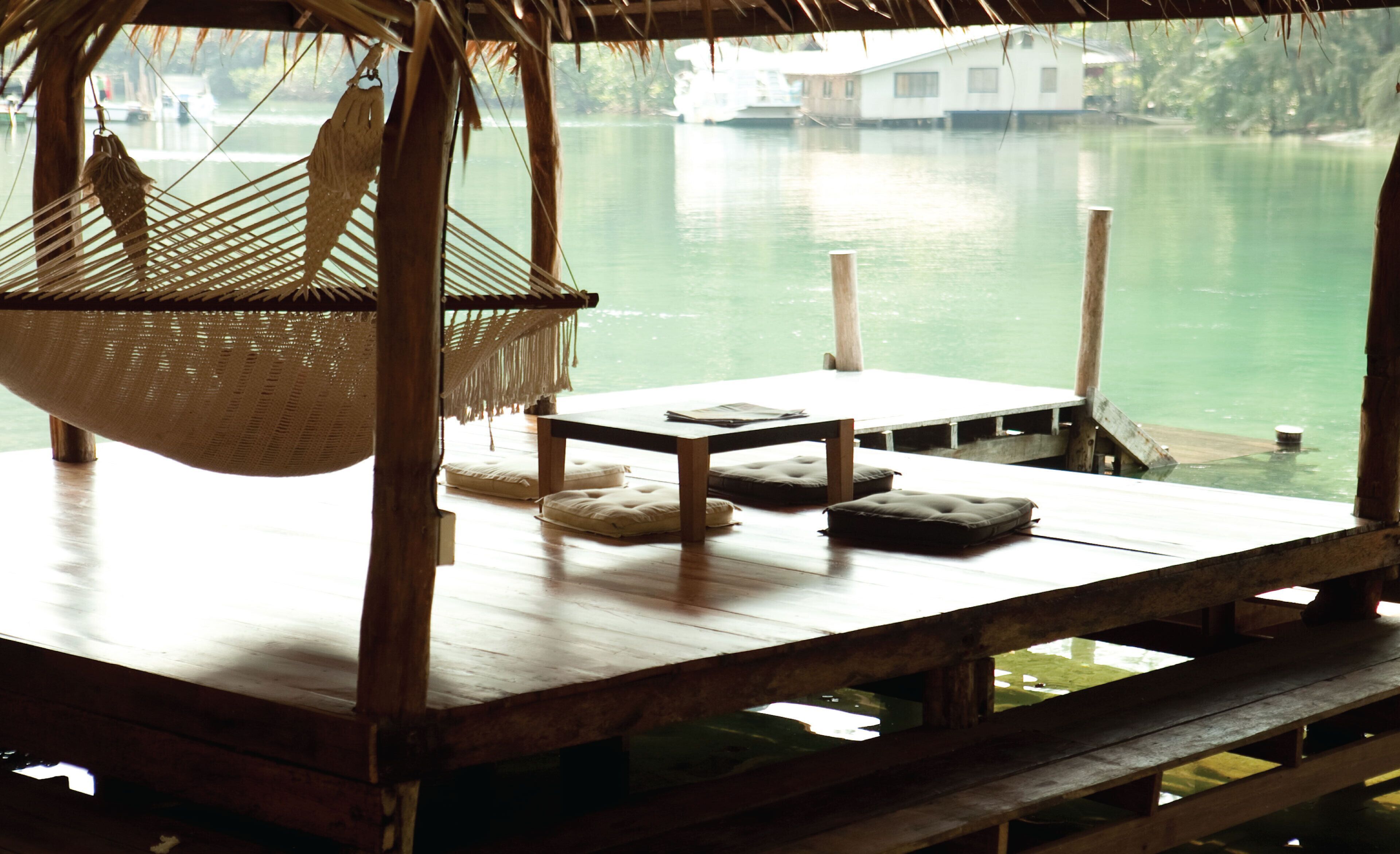Une terrasse en bois avec un hamac, des coussins et une table, le tout donnant sur une étendue d’eau calme.