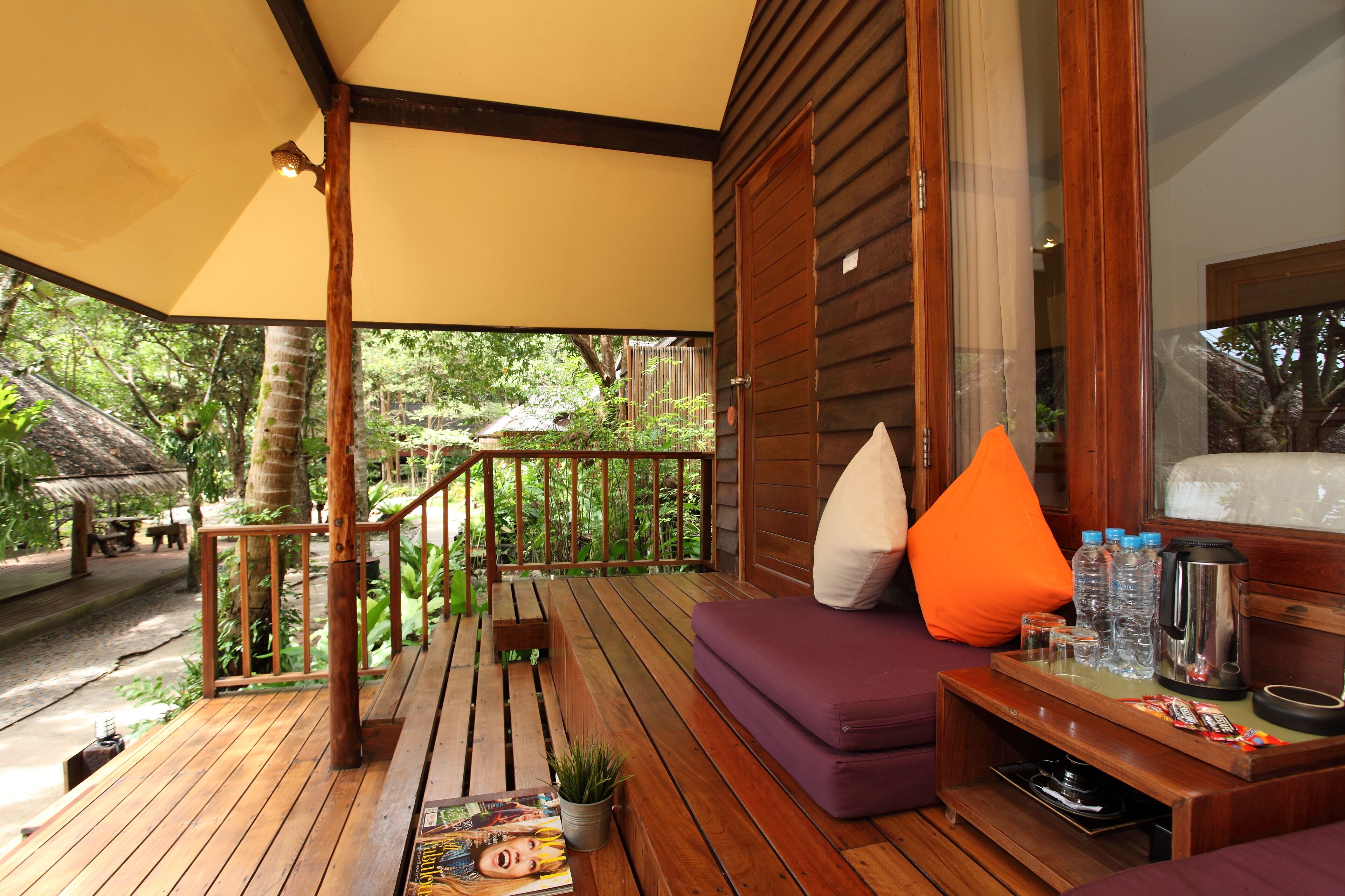 A wooden deck with a sofa and cushions, a coffee table with snacks and a kettle, and a magazine on the floor.