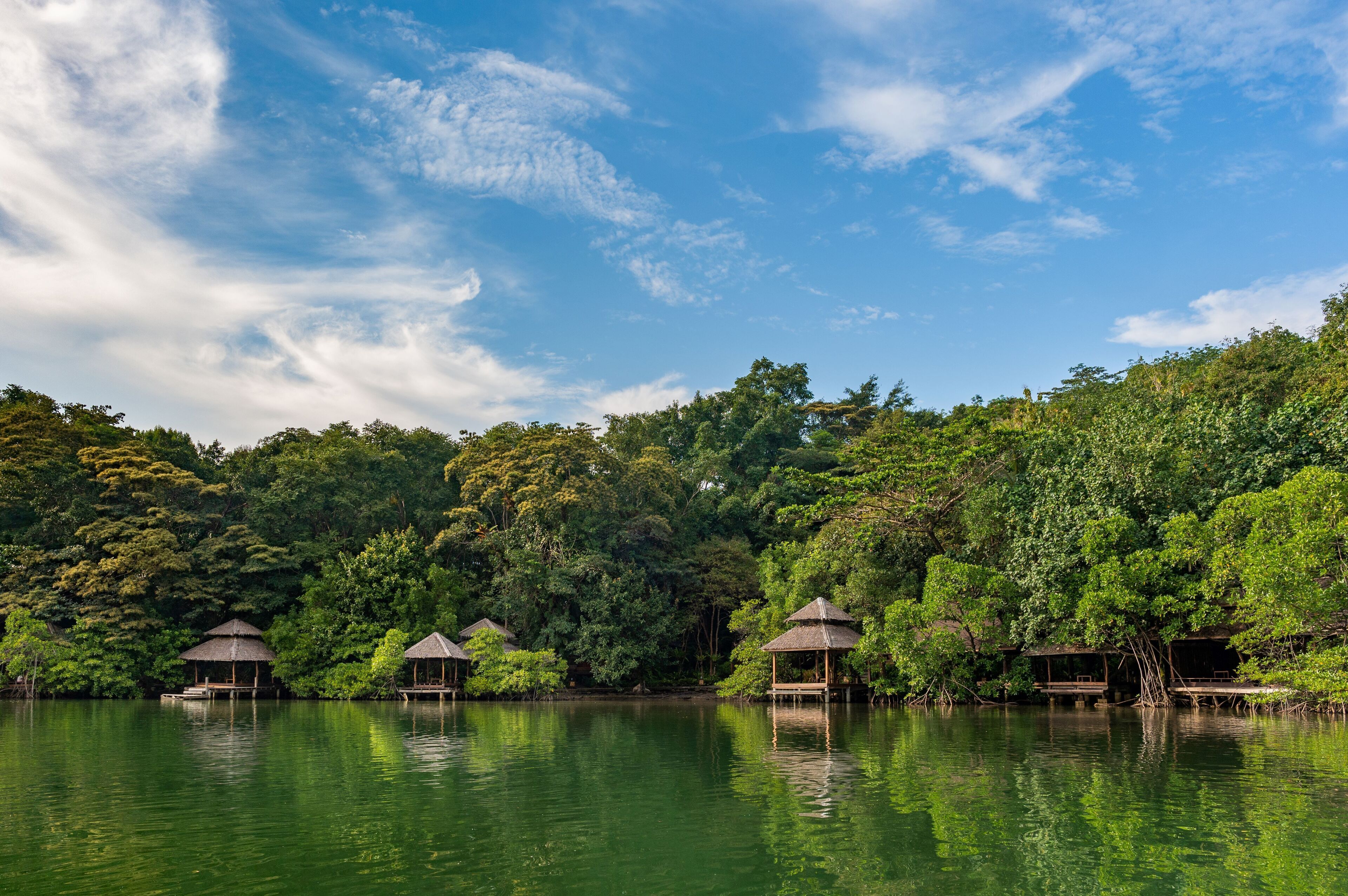 A tranquil lakeside scene with several thatched-roof huts on stilts, surrounded by dense greenery and a clear blue sky.