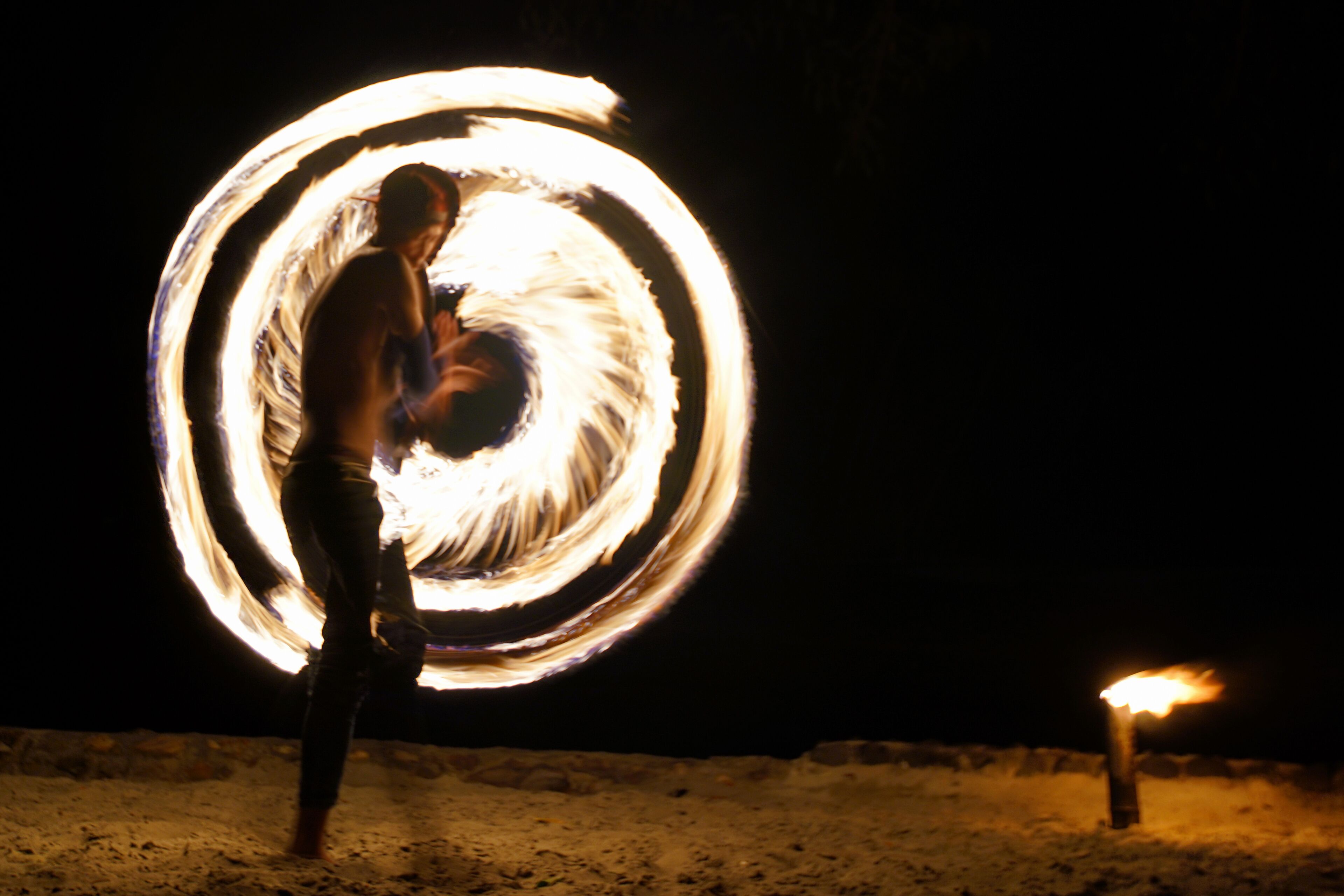 Una persona realizando una exhibición de malabares con fuego en un entorno exterior oscuro.