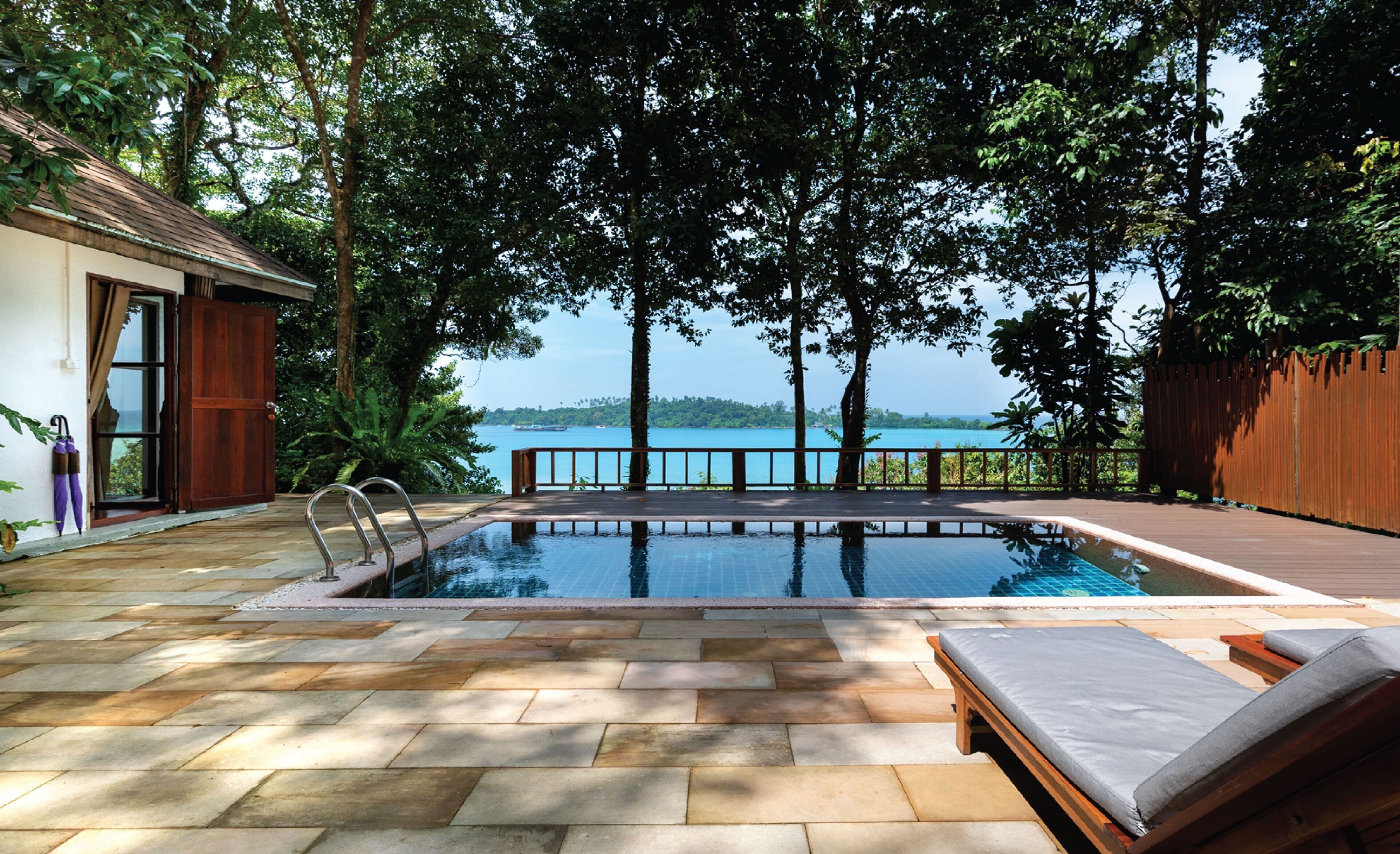 An outdoor swimming pool with a wooden deck, lounge chairs, and a view of the sea.
