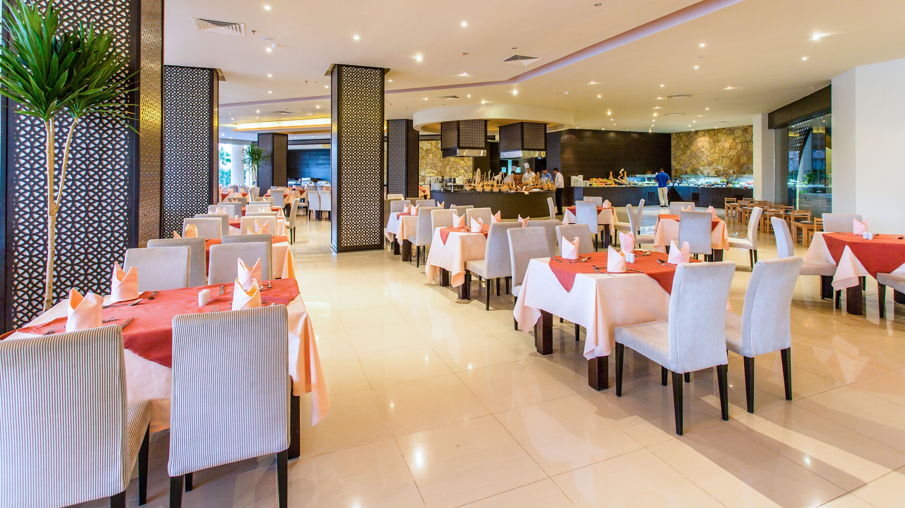 A spacious restaurant with multiple dining tables and chairs, a central bar area, and decorative plants.
