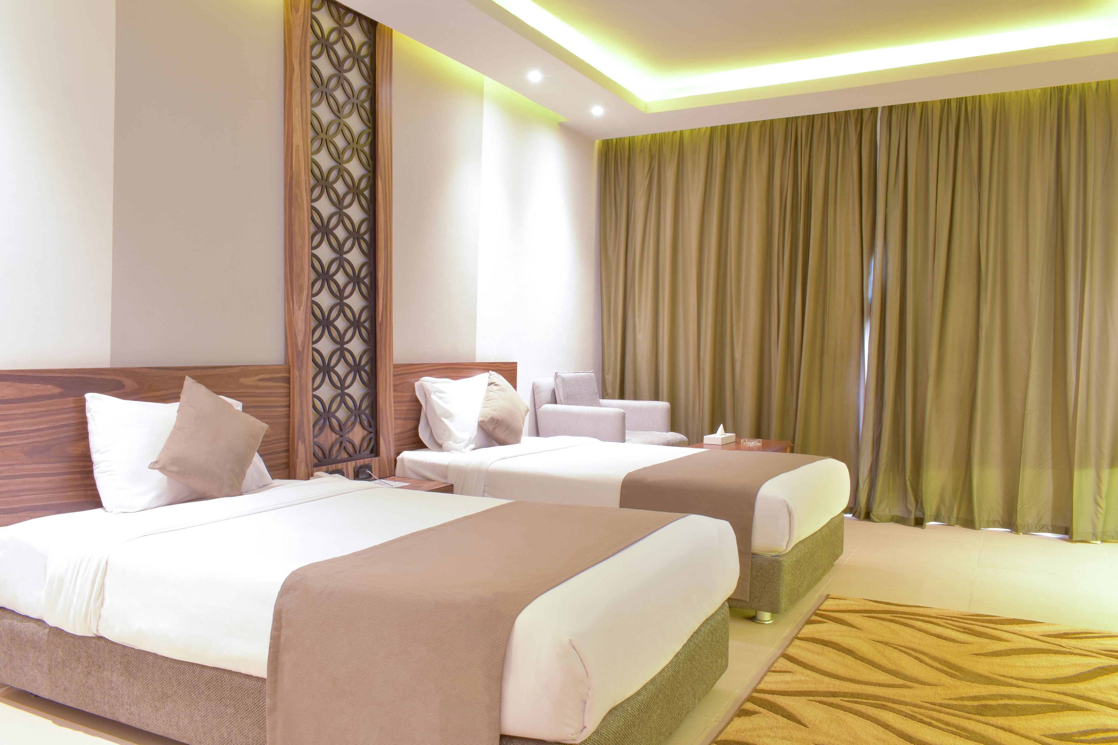 Ein Hotelzimmer mit zwei Betten, einem Holzkopfteil, einer Zierleiste, einem grünen Vorhang und einem gemusterten Teppich.
