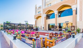 An outdoor seating area with wooden furniture and colorful cushions, in front of a building with large windows and a decorative facade.