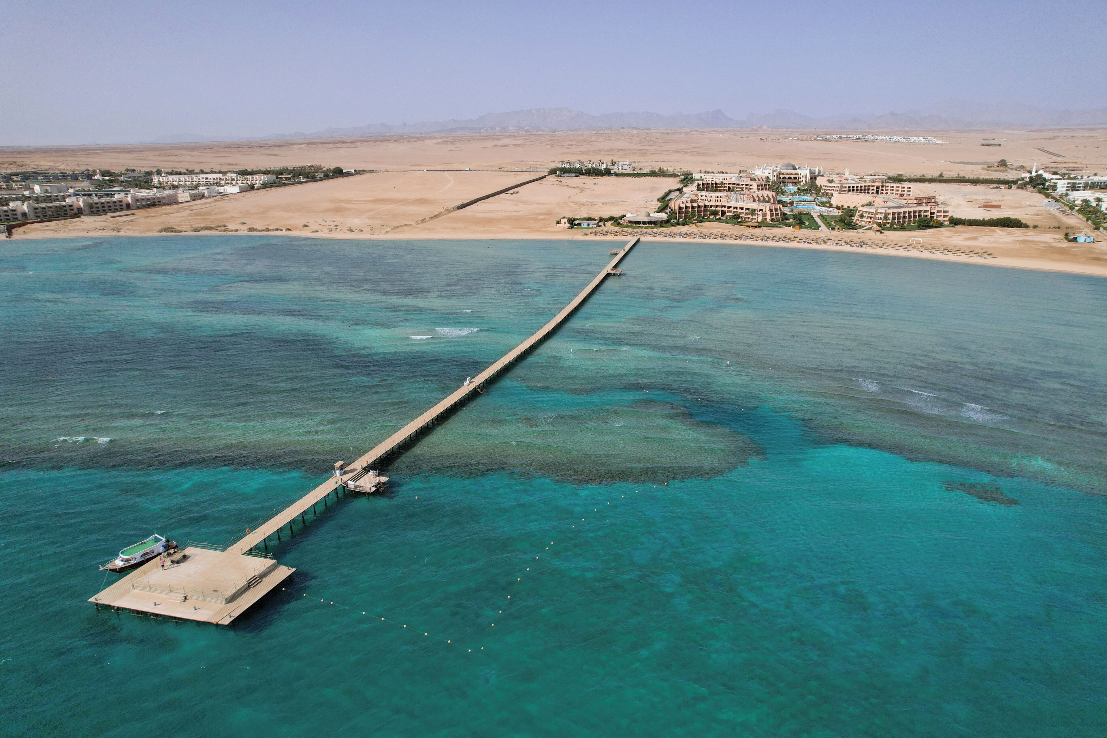 An aerial view of a coastal resort with a long pier extending into turquoise waters, surrounded by desert landscapes and hotel buildings.