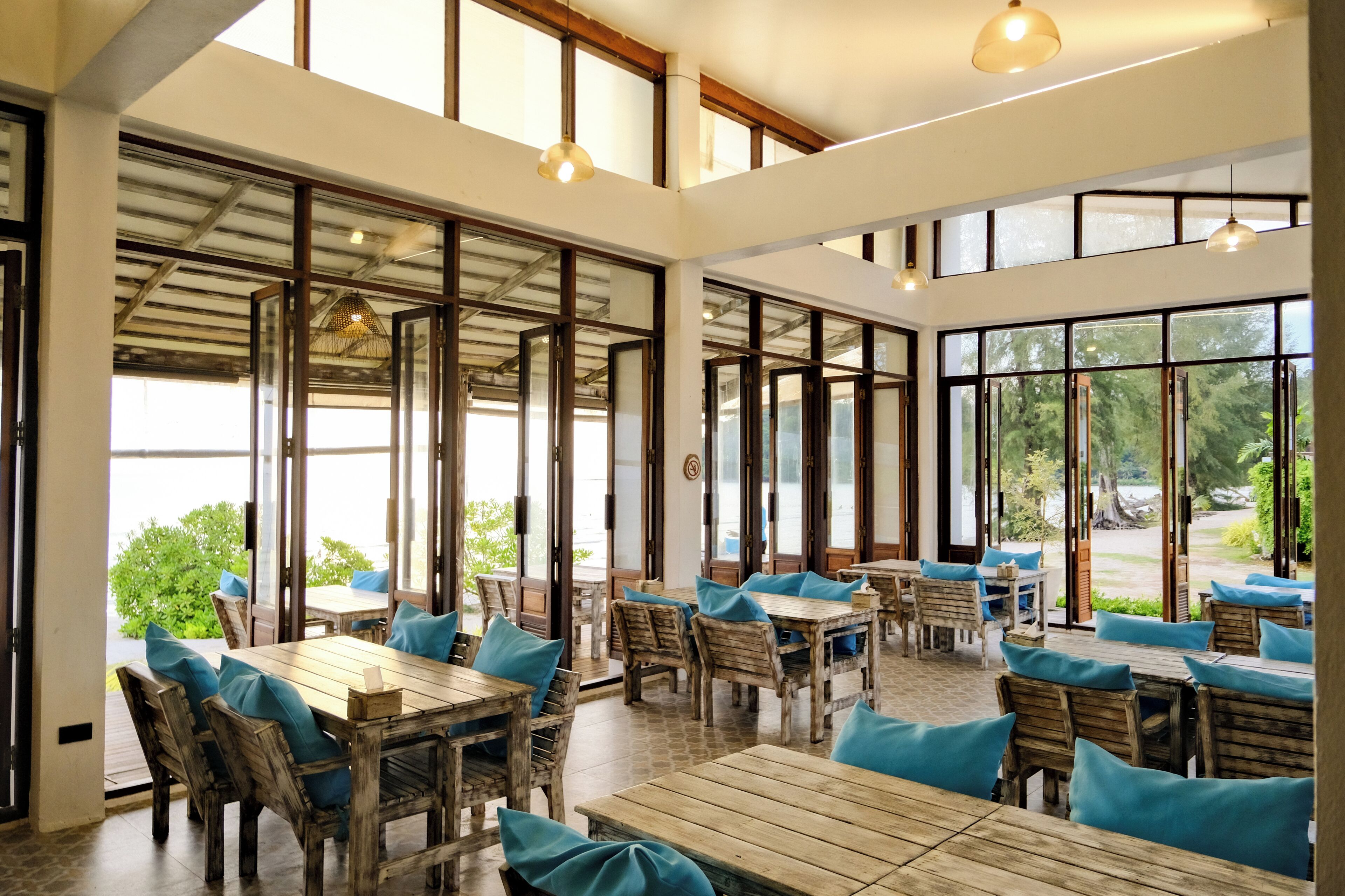 Un comedor interior espacioso con mesas y sillas de madera, ventanas amplias y lámparas colgantes.