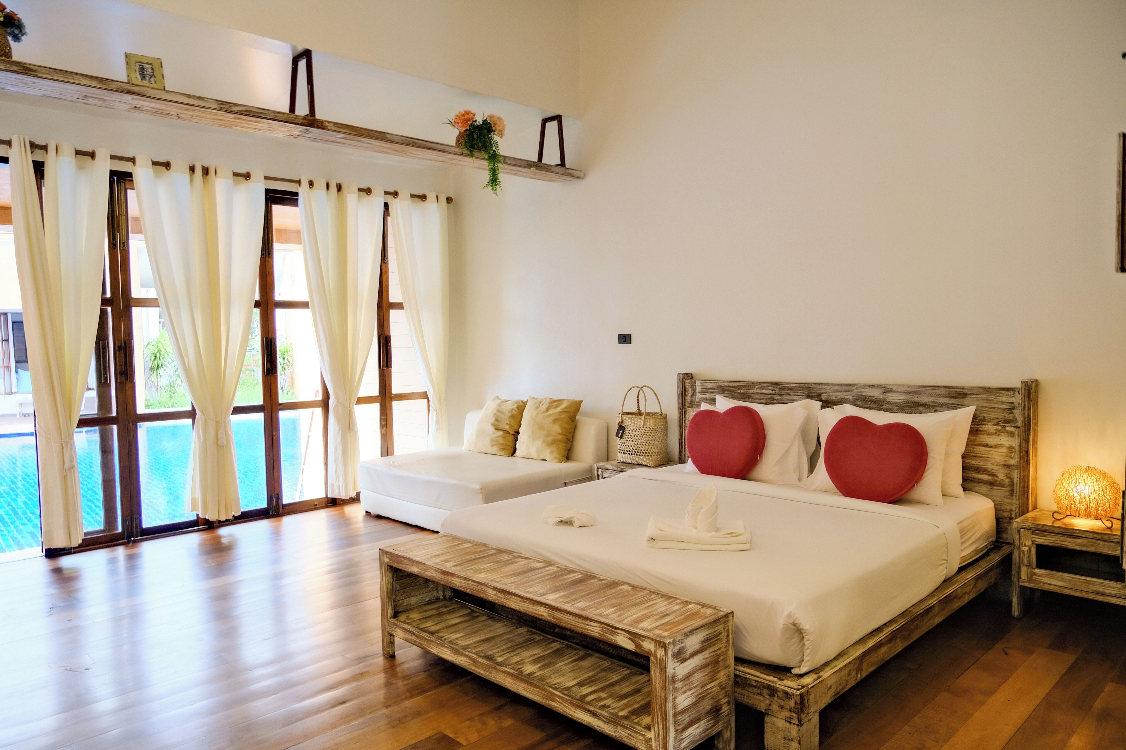 A bedroom with a wooden bed, a bench, a bedside table with a lamp, and a view of a pool.