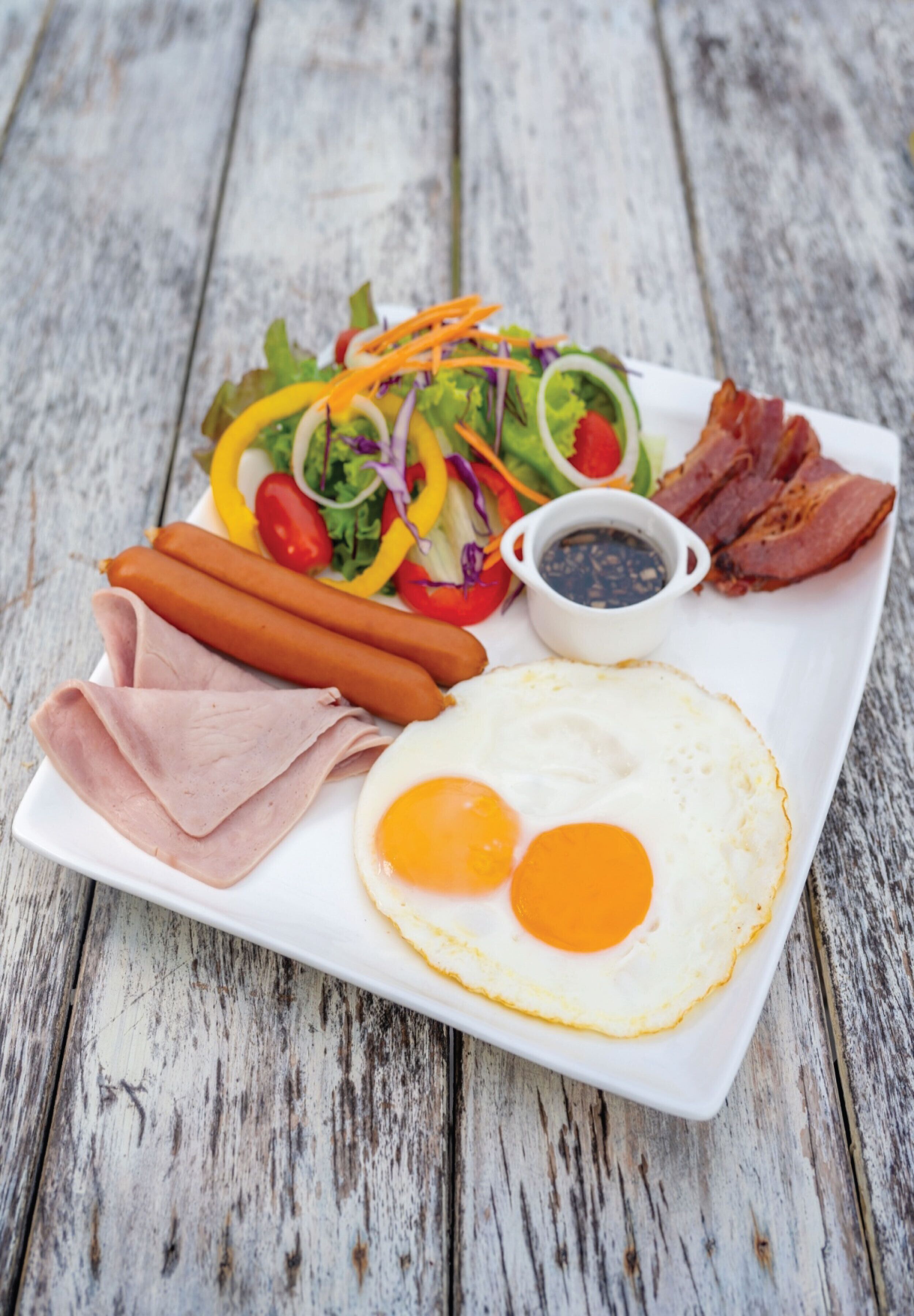 Une assiette rectangulaire blanche contenant divers aliments pour le petit-déjeuner, notamment des œufs, du bacon, des saucisses, une salade et un petit bol de sauce.