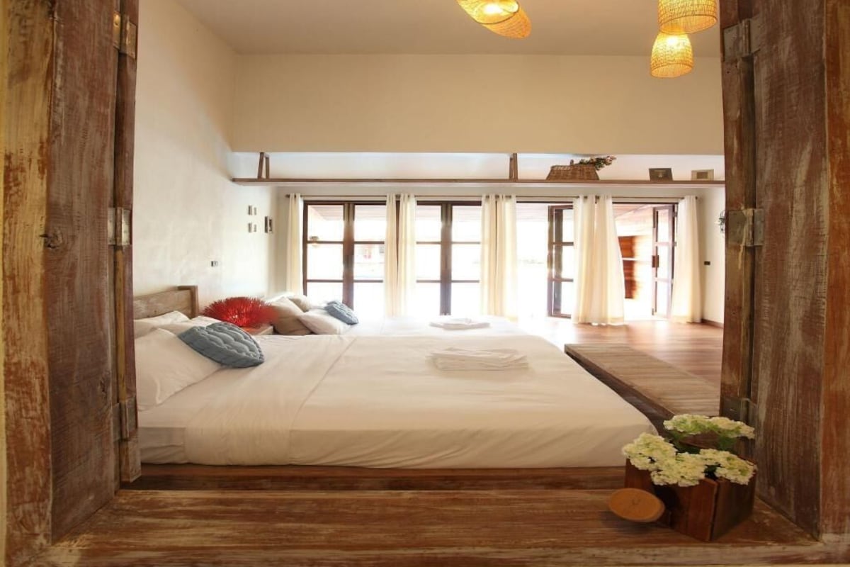 A bedroom with a large bed, wooden flooring, and a view of the outside through a door.