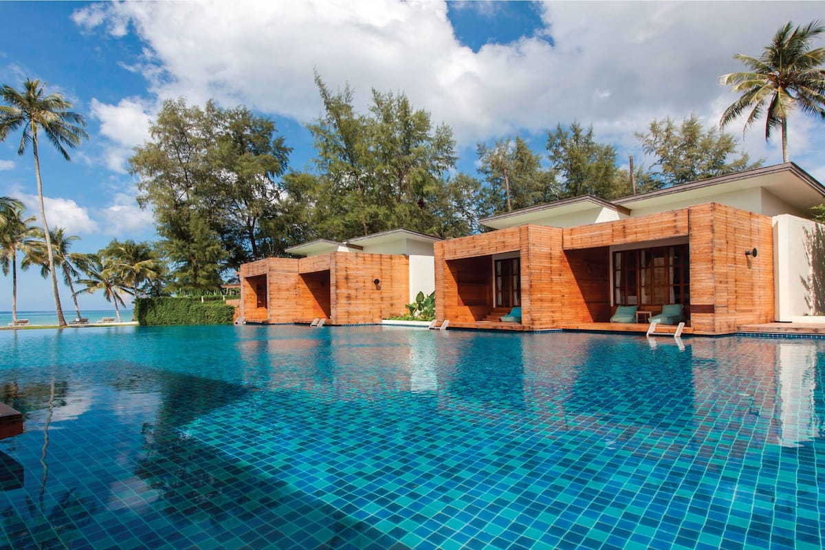 A poolside area with wooden cabanas, a clear blue pool, and palm trees.