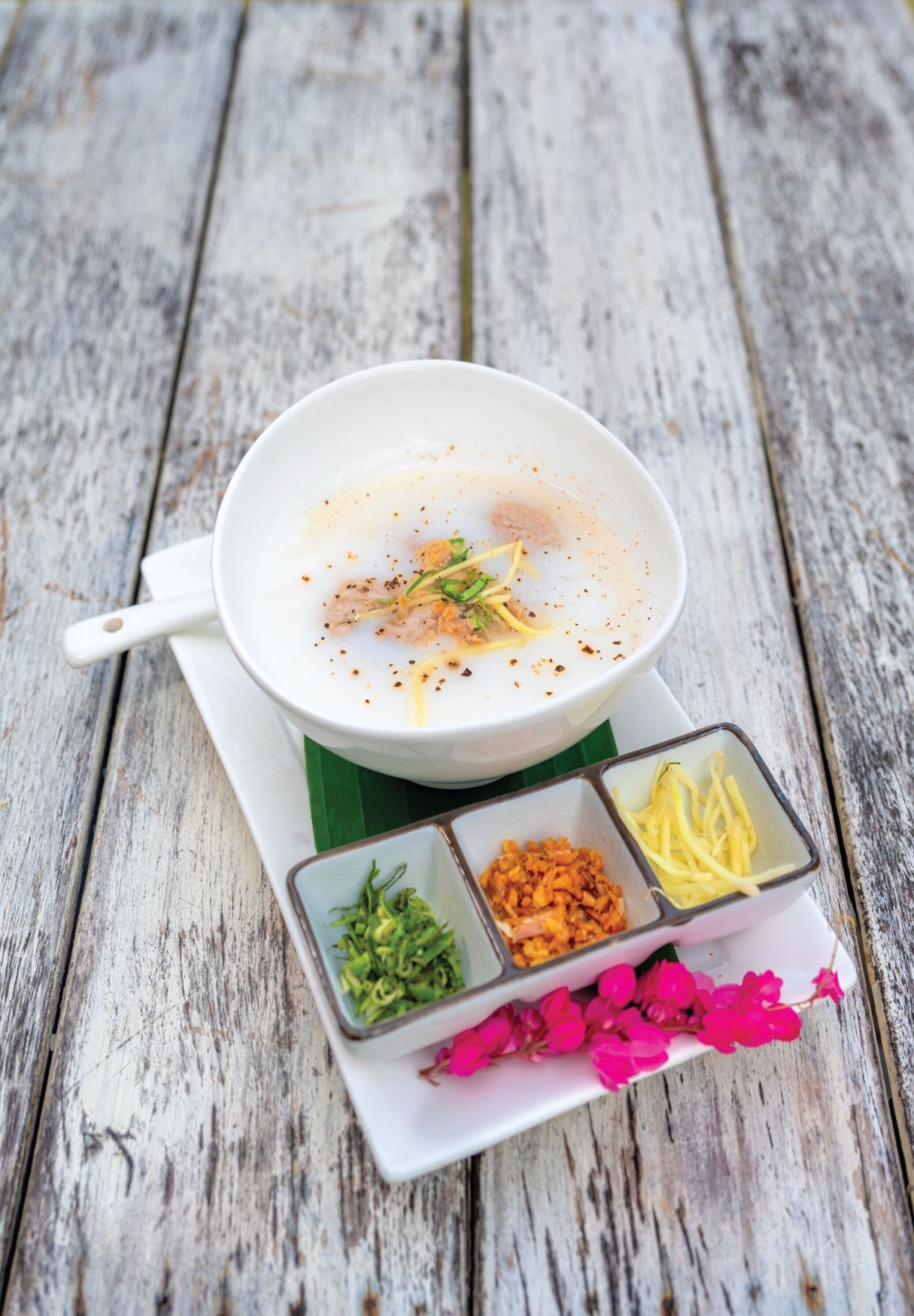 A white bowl of soup on a white tray with a green leaf underneath, accompanied by three small bowls containing different condiments.