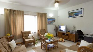 Ein Wohnzimmer mit einer Couch, zwei Sesseln, einem Couchtisch mit Blumen, einem Fernseher und einem Schreibtisch mit einem Computer.
