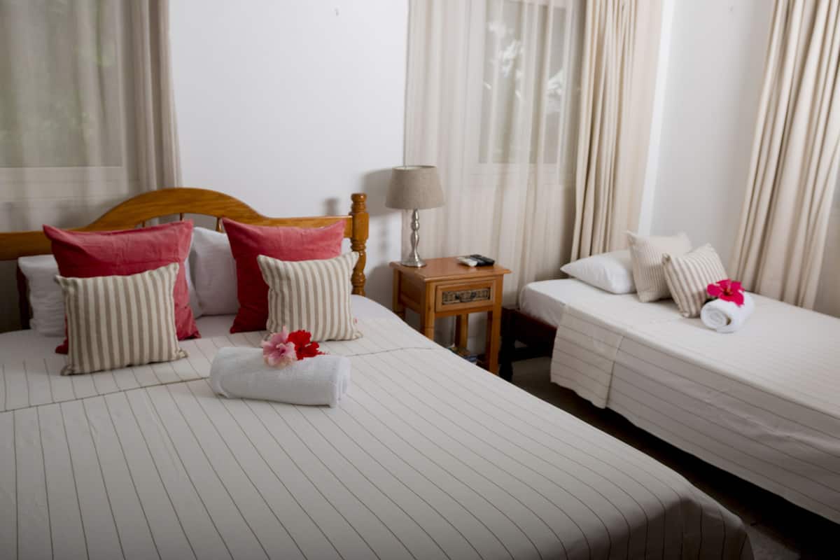 A hotel room with a large bed, two smaller beds, a nightstand with a lamp, and a window with sheer curtains.