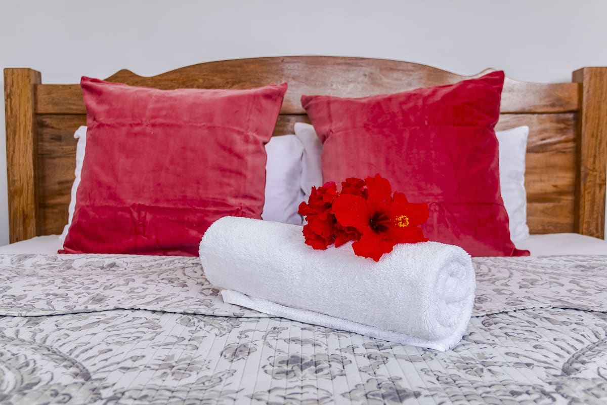 A neatly made bed with a patterned bedspread, two red pillows, a white pillow, and a rolled white towel with red flowers.