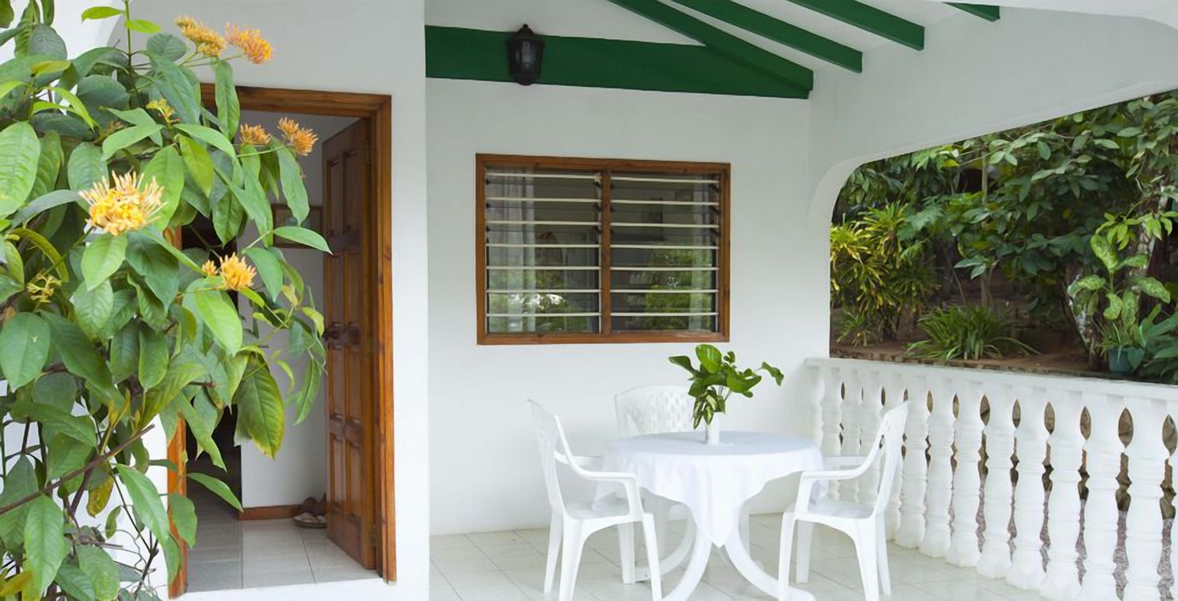 Une terrasse avec une table et des chaises blanches, une porte en bois, une fenêtre avec des stores et un mur végétal.