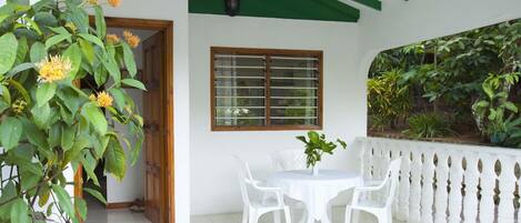 A patio with a white table and chairs, a wooden door, a window with blinds, and a green wall with plants.