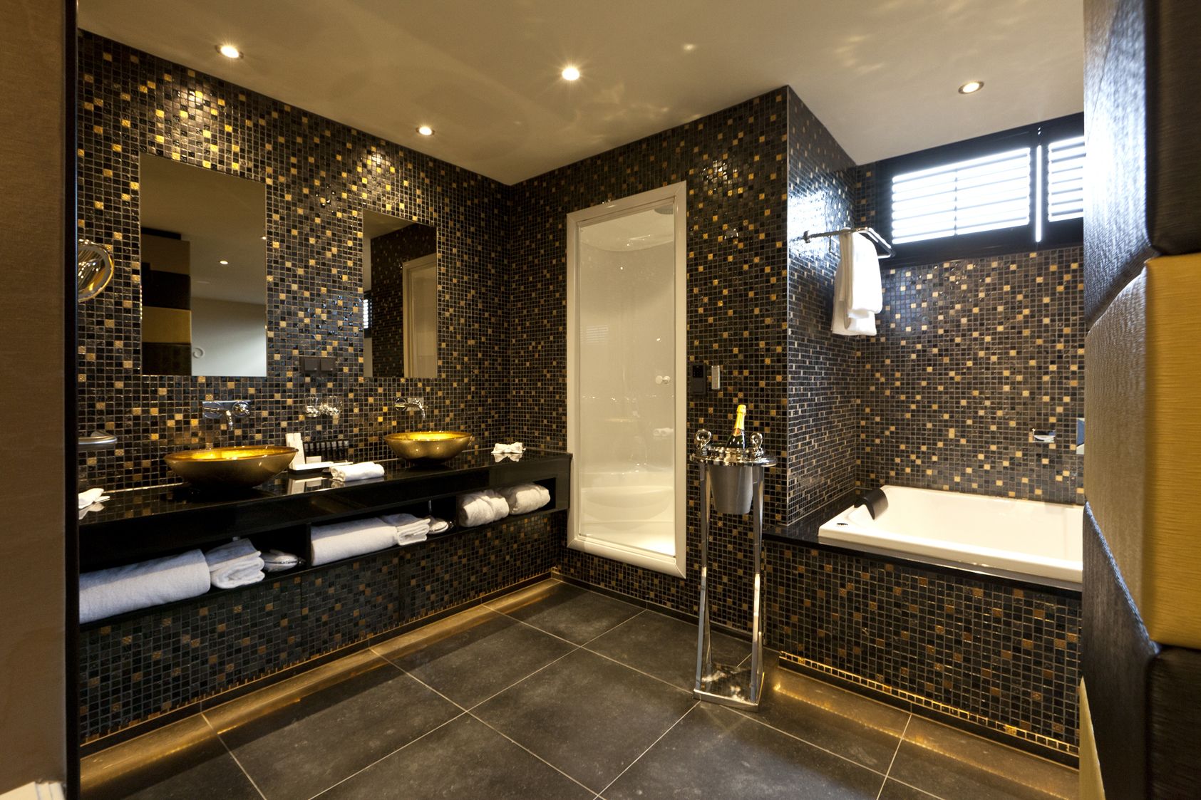 Un baño moderno con un salpicadero de mosaico negro y dorado, un lavamanos grande de color dorado, una ducha con mampara de vidrio y una bañera blanca.