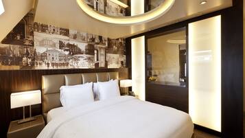 A modern hotel room with a large bed, a headboard with a cityscape mural, a round ceiling light, and a large mirror reflecting the room.