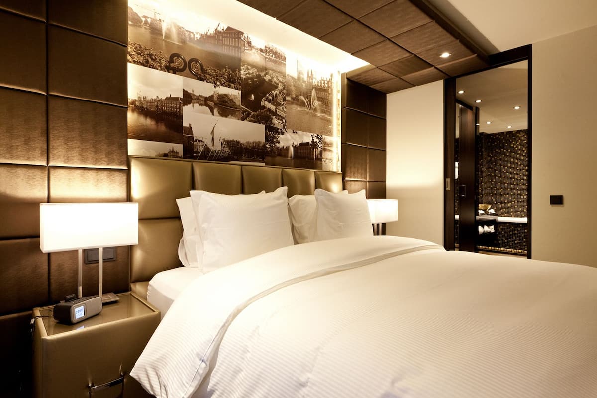 Une chambre d’hôtel moderne avec un grand lit, deux lampes de chevet, une table de chevet avec une horloge et une œuvre d’art accrochée au mur.
