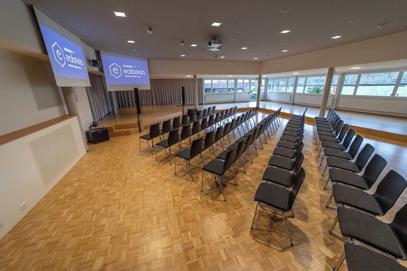 A conference room with rows of chairs, a stage, and a large screen displaying the Eckstein logo.