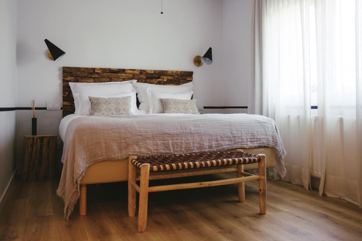 A bedroom with a wooden headboard, a bed with white linens, a bench at the foot of the bed, and a window with sheer curtains.